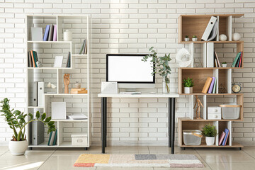 Workplace with computer, plant branches in vase and shelving units near white brick wall