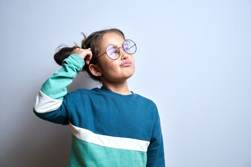 Funny little girl in glasses scratching her head doubts, makes decision isolated on yellow studio...