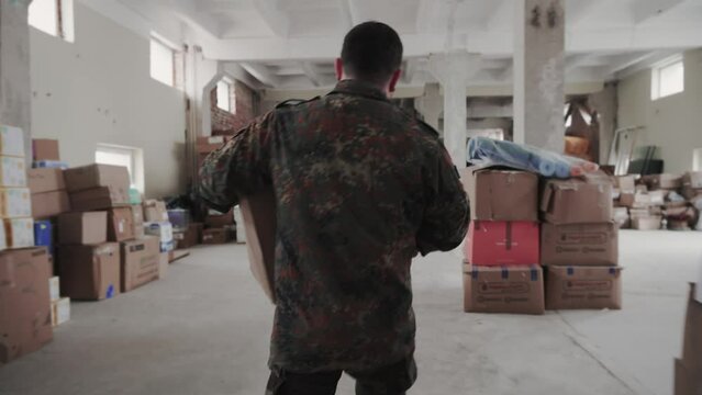 Military man carries a heavy cardboard package and puts it on the pile of boxes in the humanitarian war centre.