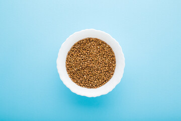 White bowl with dry brown buckwheat on light blue table background. Pastel color. Closeup. Healthy food. Top down view.