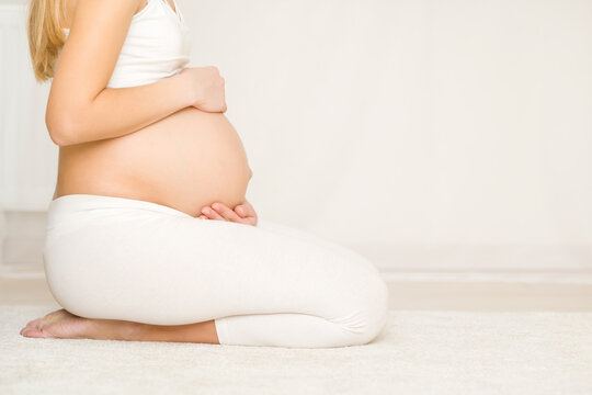 Young adult pregnant woman in white clothes sitting on home carpet. Hand touching big naked belly. Pregnancy concept. Expectation time. Empty place for text on light gray wall background. Side view.