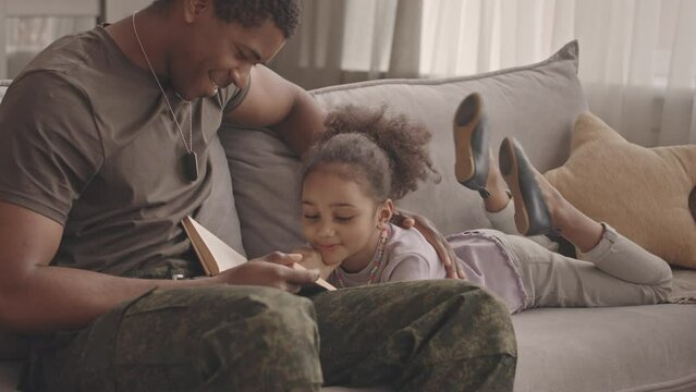 Slowmo of cute 6 year old African American girl lying on her stomach on sofa reading book aloud while spending leisure time together with her loving father in military uniform