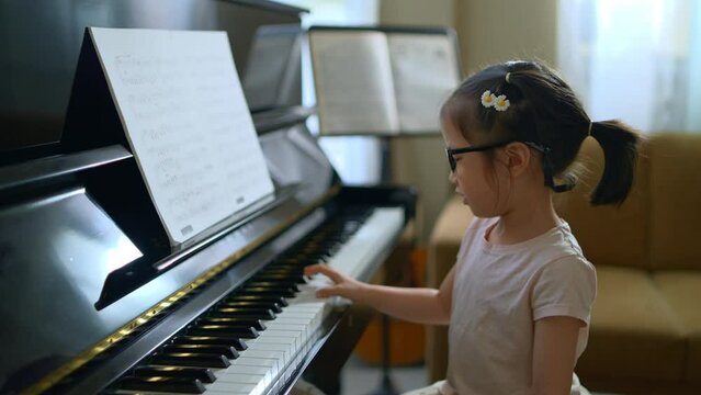 Asian toddler girl look at camera with smiling while playing piano, exploring music instrument, performance, early childhood music education, leisure activity, side view girl kid playing piano.