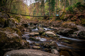 Moss stones in the river with suspension bridge in a mountain valley and green forest. Natural landscape torrent Buchberger Leite in the Bavarian Forest, Germany.