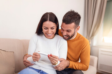 Enamoured couple finding out results of a pregnancy test sitting on the bed. Happy couple checking pregnancy test sitting on a couch in the living room at home. Family, parenting and medical concept