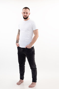 Young barefoot bearded man with short dark hair in white T-shirt, black jeans, standing, thrusting hands into pockets.