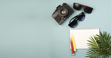 A large horizontal banner with copy space, an empty notebook with white sheets, a camera, colored markers, glasses and palm leaves on a blue background. The concept of travel, leisure, freelance and b