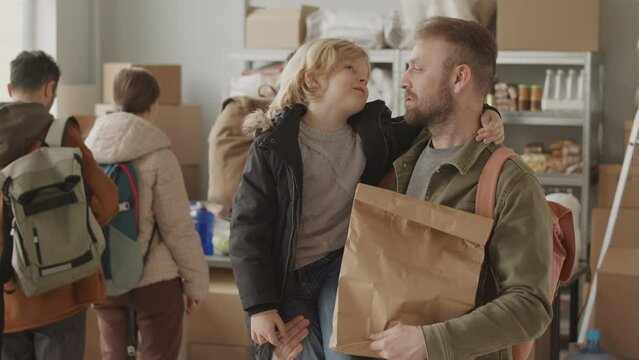 Medium slowmo portrait of single father and son talking then looking at camera standing indoors at food bank where refugees and people in need can get basic provisions