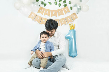 Naklejka premium Asian baby boy celebrating first birthday,A father sits and hug his son smiling on a floor with a minimal background,white balloons and birthday flag.Selective focus