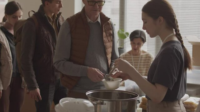 Medium slowmo of young female volunteer pouring hot soup in disposable bowls to refugees and homeless people standing in queue at local food bank