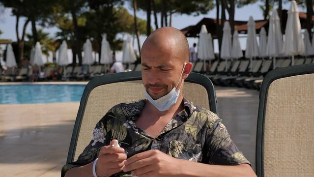 Portrait of a young sick man with symptoms of coronavirus in a hotel on vacation, he sits on a chaise longue by the pool, sprays his nose with spray and puts a mask on his face.