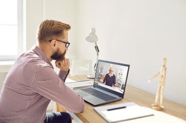 Serious focused young man in glasses sitting at working desk with laptop computer, notepad and pen at home, watching educational video lecture or having online class with experienced senior teacher