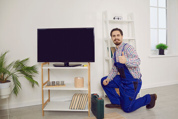 Fototapeta na wymiar Happy repairman with toolbox, in blue overall uniform showing thumbs up sign gesture after he's checked, fixed or installed LED LCD widescreen television set in living room. Warranty TV repair concept