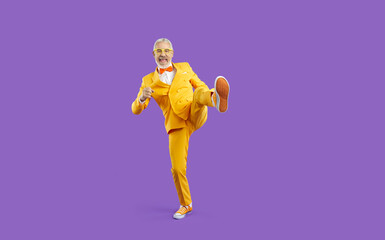 Cheerful eccentric and funny senior man in yellow suit isolated on purple background. Stylish cool retiree in bright suit and sneakers makes big wide step while having fun in studio. Full length.