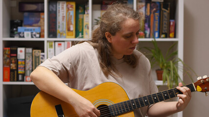 A girl plays an overdub on an acoustic guitar sitting in a room. Behind her is a rack of board...