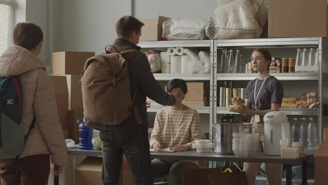 Slowmo of female volunteers from npo providing food supplies to homeless, refugee or needy people of different age, standing in queue indoors