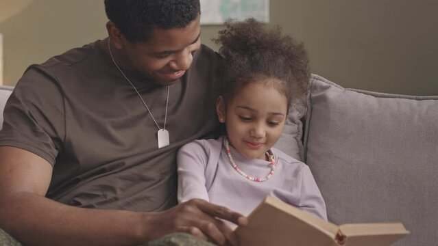 Waist up slowmo of smiling young Black man in military uniform and his pretty little daughter reading book together while sitting on couch at cozy home