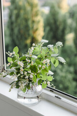 Bouquet of cut jasmine branches in a glass vase on the windowsill of the glazed loggia with a view of the city park.