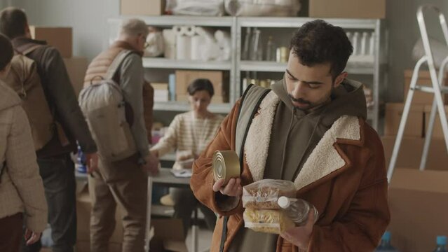Slowmo portrait of bearded refugee man with food and water supplies in hands looking at camera sadly while standing indoors where volunteers providing help and assistance to asylum seekers