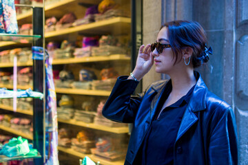 headshot of a beautiful woman of latin features with one hand raised holding her sunglasses observing merchandise outside store