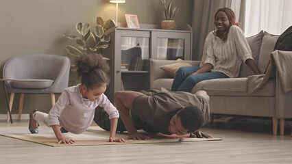 Slowmo of fit African American soldier in military uniform and his cute 6 year old daughter doing pushups together on rug in living room and his wife and girls mom watching and smiling