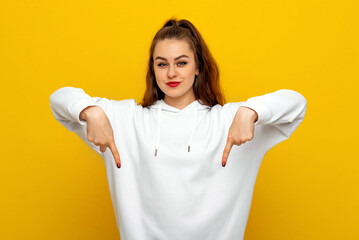 Beautiful brunette girl in white casual style sweatshirt standing against yellow background and pointing fingers down, showing advertisement. Young emotional woman
