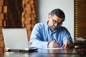 Fototapeta premium Positive senior bearded man with grey hair drinking coffee and using laptop at cafe, copy space. Stylish aged businessman in burgundy jacket enjoying his tea while working online.