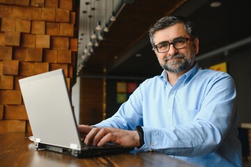Mature businessman drinking coffee in cafe. Portrait of handsome man wearing stylish eyeglasses using laptop, looking at camera, smiling. Coffee break concept