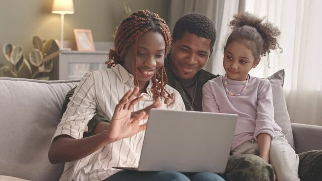 Medium slowmo of happy African American military family of three talking with friends or relatives through video chat on laptop, sitting together on couch in living room
