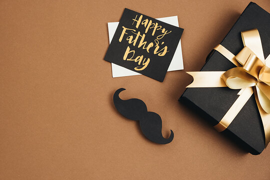 Horizontal images of handmade black gift box, card with phrase happy father's day and retro stylish black paper photo booth props moustaches on brown studio background wall. Holiday concept