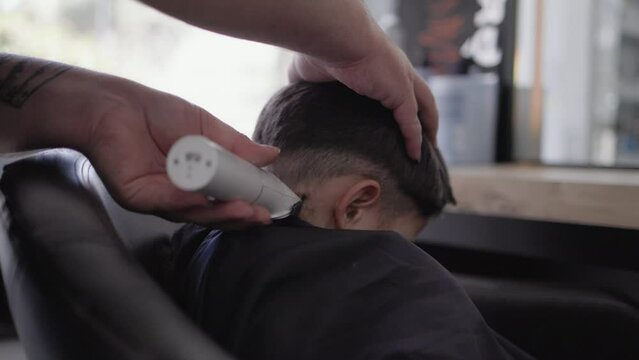 The hairdresser cuts the little boy's hair with a clipper holding his head. Child in salon