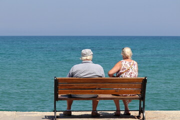 Elderly couple sitting on a wooden bench on blue sea background, rear view. Old woman and man on a beach, summer travel and enjoying life in retirement