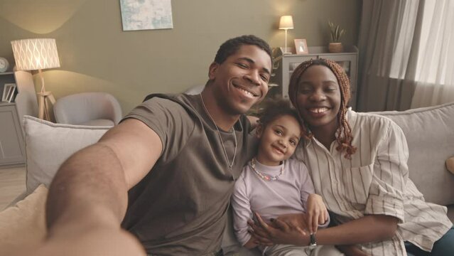 Handheld POV slowmo of delighted African American soldier smiling at camera while taking selfie with his beloved wife and pretty little daughter, sitting together on soft grey couch in living room - Powered by Adobe