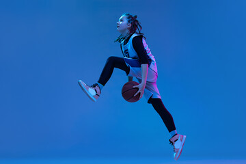 Fototapeta na wymiar Dynamic portrait of teen girl, basketball player training, jumping with ball isolated over blue studio background in neon. Defender