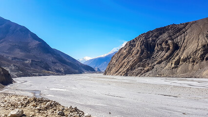A view on a dry bottom of Himalayan valley. The valley is located in Mustang region, Annapurna Circuit Trek in Nepal. In the back there is snow capped Dhaulagiri I. Barren and steep slopes