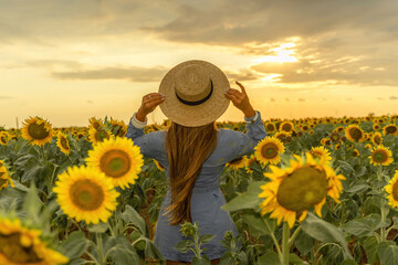 Beautiful middle aged woman looks good in a hat enjoying nature in a field of sunflowers at sunset. Summer. Attractive brunette with long healthy hair.