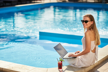 Self employed woman doing remote work with laptop sitting near swimming pool at tropical spa with...