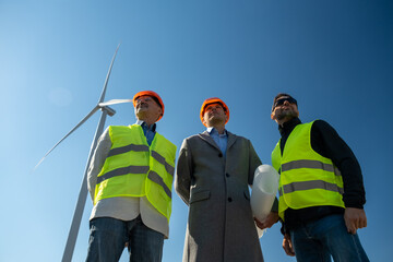 Wind turbine rotates behind senior engineer and technicians with project paper. Specialists look at built stations of rotating windmills low angle shot