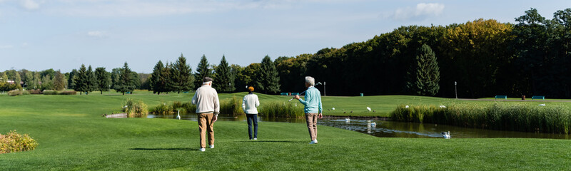 Obraz premium back view of senior multiethnic men with golf clubs walking near pond on green lawn, banner.