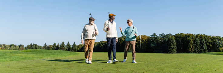 senior interracial friends walking with golf clubs on field, banner.