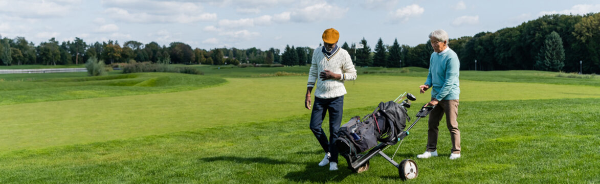 Asian Senior Man Walking With Golf Cart Near Wealthy African American Friend, Banner.