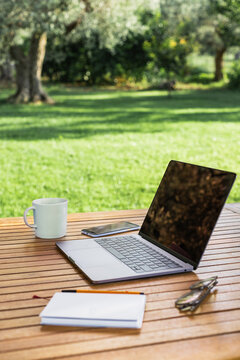 Computer, tazza, quaderno e occhiali su tavolo di legno in un giardino d'estate 