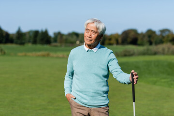 asian senior man standing with hand in pocket and holding golf club on green field.