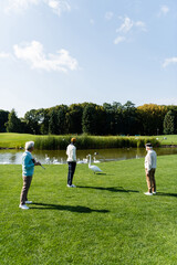 back view of senior multiethnic men with golf clubs standing on green lawn near swans.