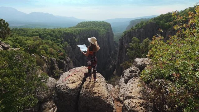 Back view of young woman in summer outfit and straw hat standing on high rock and holding paper map in hands. Female traveler discovering new interesting places during vacation in Turkey.
