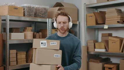 Overjoyed postman wearing headphones listening music while carrying parcels and sitting at his workplace with laptop at the table.