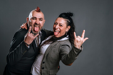 punk-style man and woman wearing jacket suits pose in studio shot looking at camera, sticking out their tongues and making horns with their hands with cheerful and casual expression