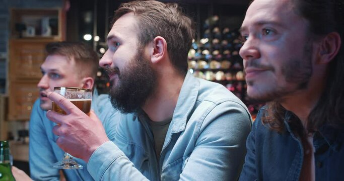 Group of young men hang out in bar, drinking beer and discussing TV broadcast. Three friends having nice conversation, smiling, nodding and having good time.