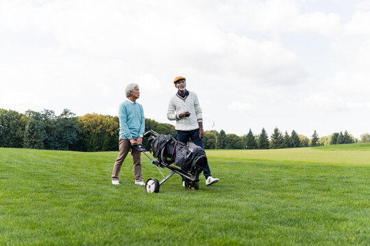 asian senior man walking with golf cart near african american friend.