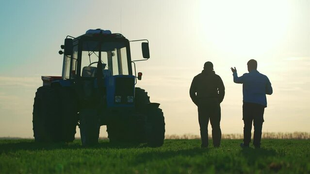 Teamwork concept. Silhouette two male farmers walking in a green field against sunset. Team farmers stand in a field on the background of agricultural machinery. Agronomists discuss harvest.